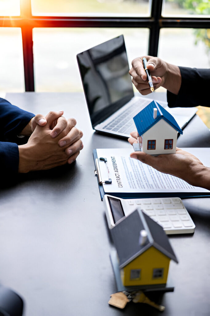 Two people at a table with a house model, keys, and a contract, suggesting a real estate transaction.