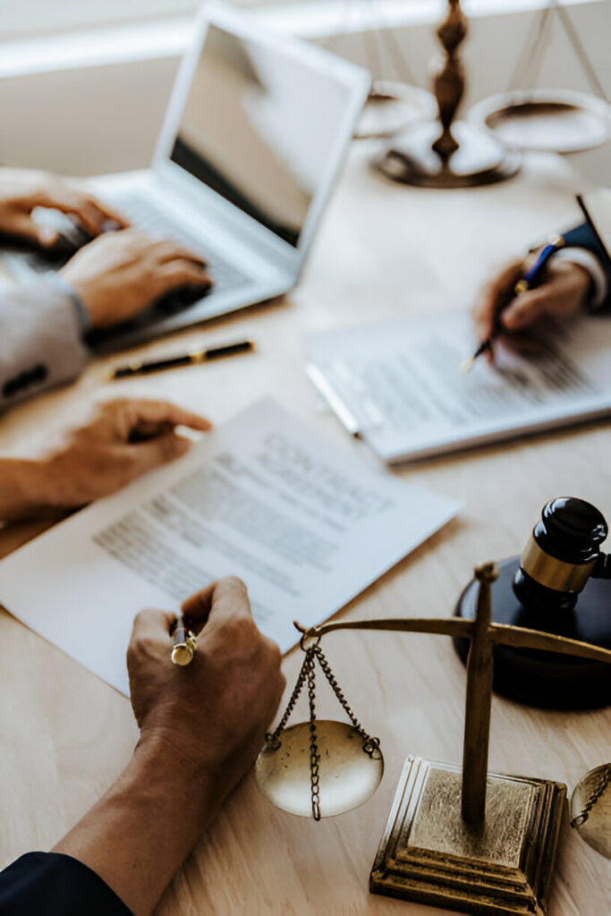 Hands signing a legal document on a desk with a laptop, scale of justice, and gavel in the background.